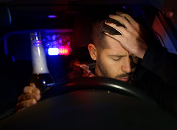 A photo of a man sitting behind his car's steering wheel and holding a bottle of beer while pulled over by the police.